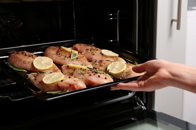 Photo of Woman putting chicken breasts with lemon and rosemary into oven, closeup