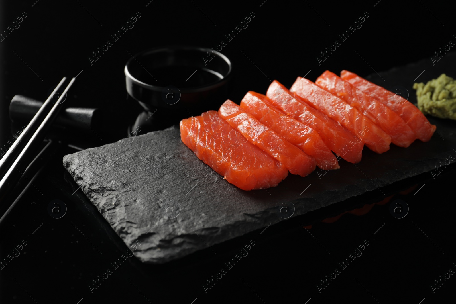 Photo of Delicious salmon sashimi, wasabi, soy sauce and chopsticks on black mirror surface