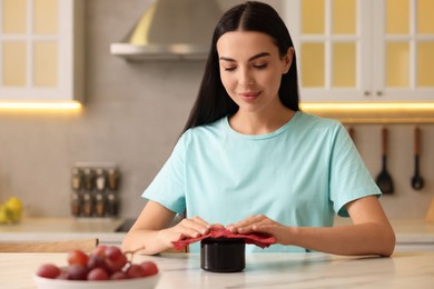 Photo of Young woman packing jar of jam into beeswax food wrap at table in kitchen