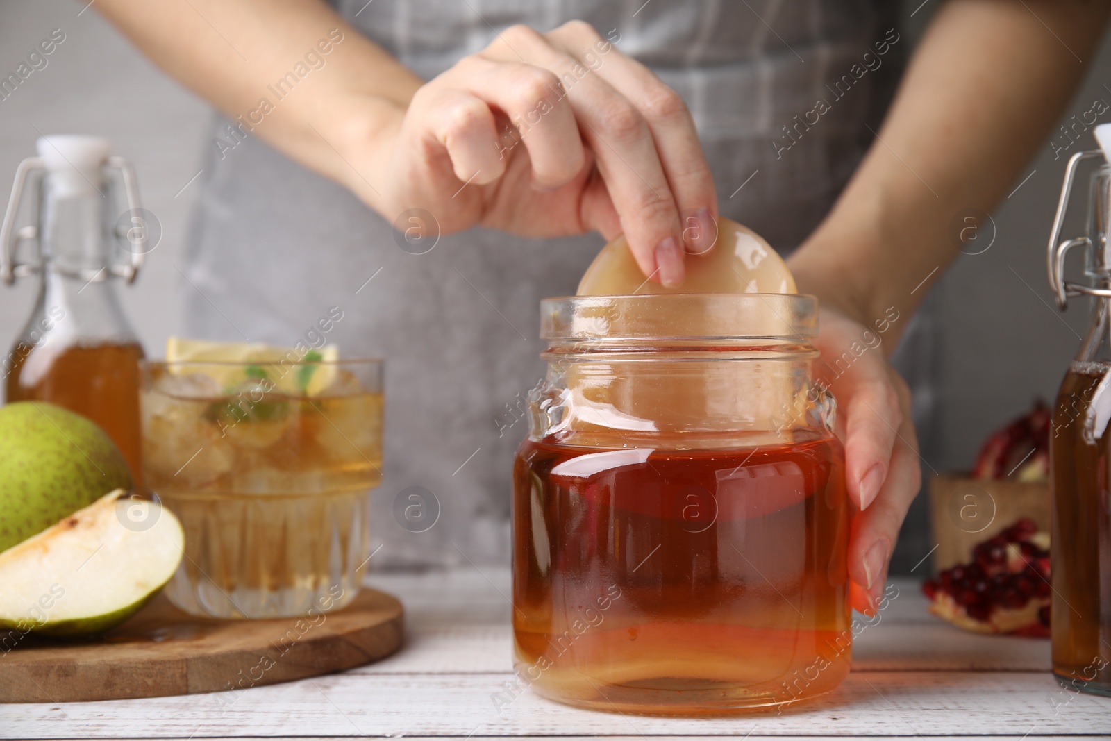 Photo of Woman putting Scoby fungus into jar with kombucha at white wooden table, closeup