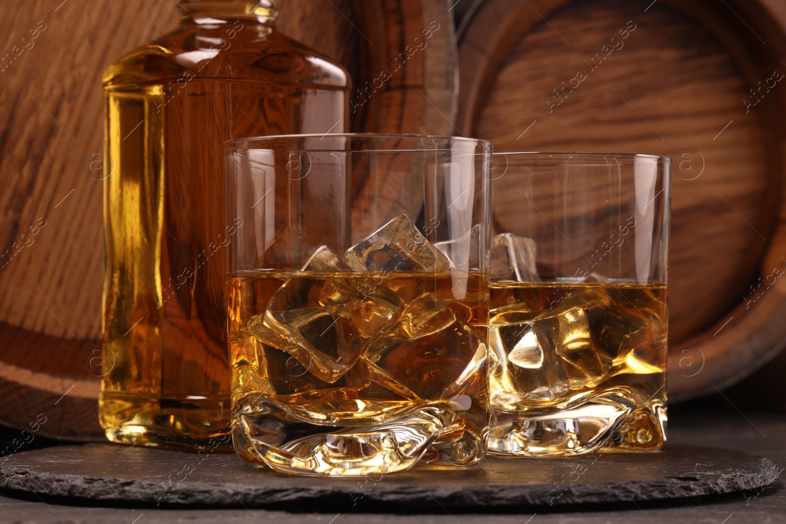 Photo of Whiskey with ice cubes in glasses, bottle and wooden barrels on black table