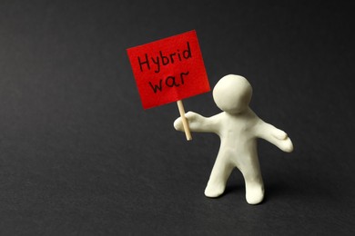 Photo of Human figure made of white plasticine holding sign with words Hybrid War on black background