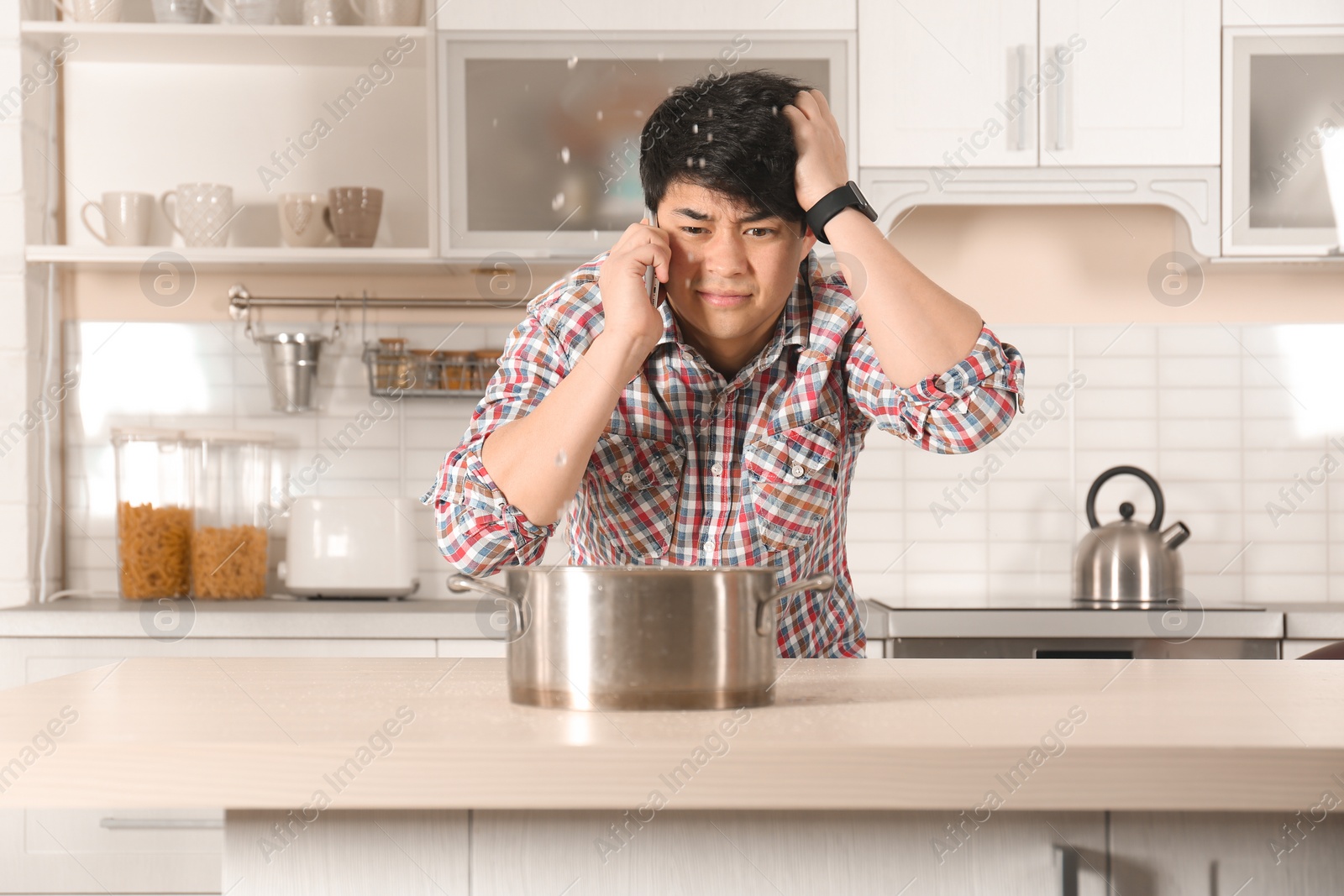 Photo of Emotional man calling plumber near table with saucepan under leaking water from ceiling in kitchen