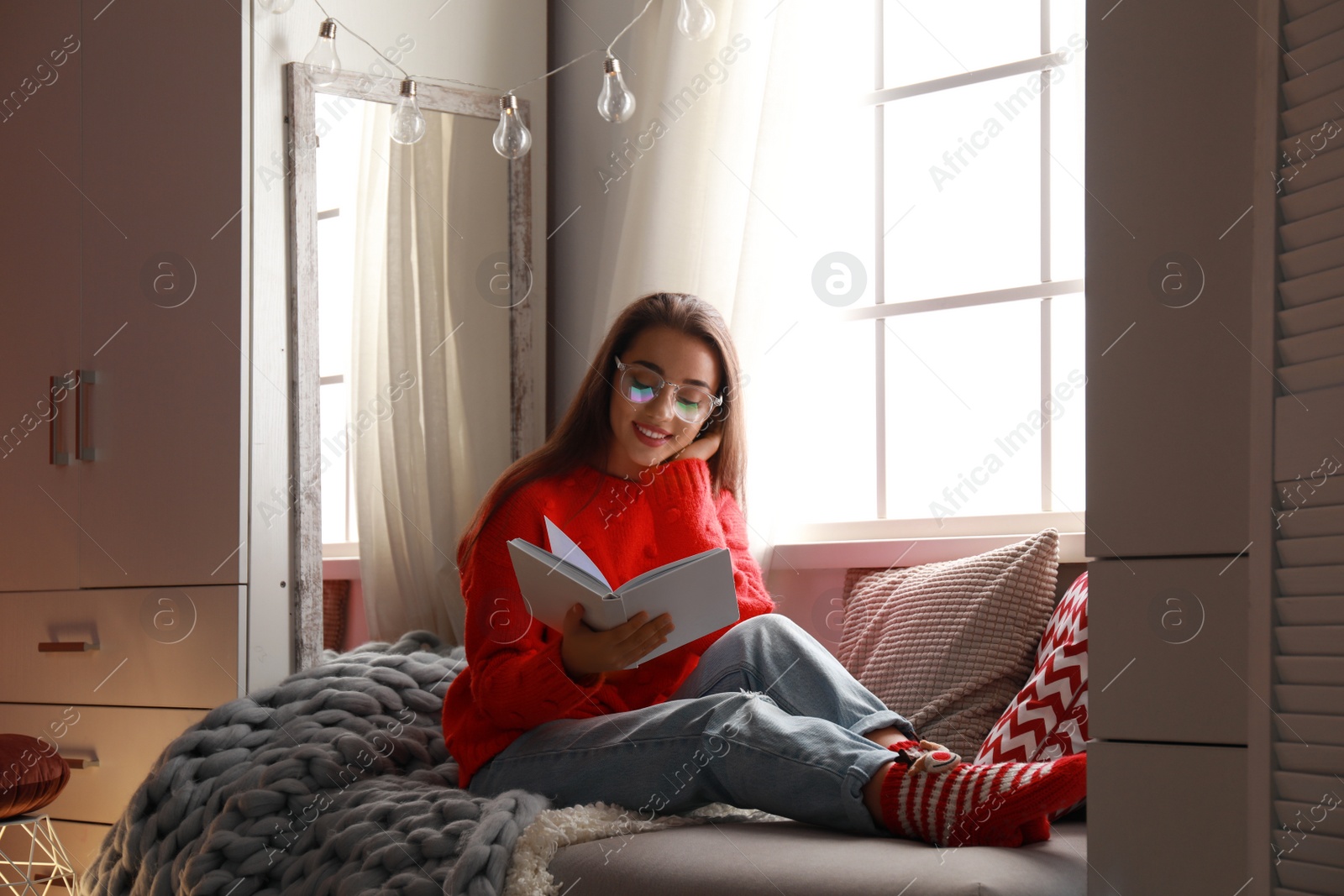 Photo of Young woman reading book near window at home. Winter season