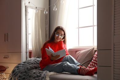 Photo of Young woman reading book near window at home. Winter season