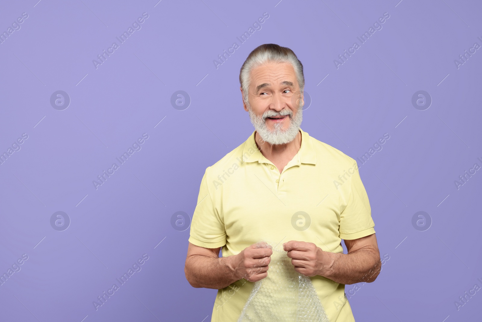 Photo of Happy senior man popping bubble wrap on light purple background, space for text. Stress relief