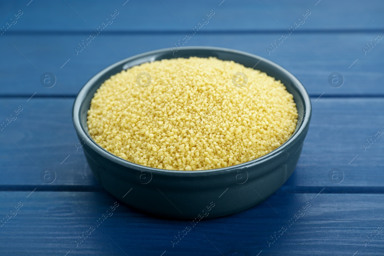 Photo of Bowl of raw couscous on blue wooden table, closeup