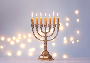 Photo of Golden menorah with burning candles against light grey background and blurred festive lights