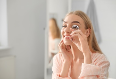 Photo of Young woman curling her eyelashes near mirror indoors