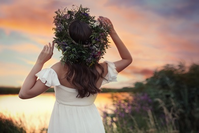 Photo of Young woman wearing wreath made of beautiful flowers outdoors at sunset, back view