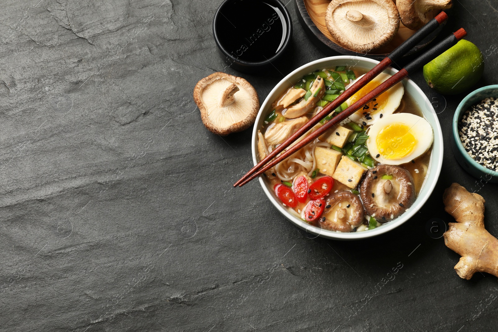 Photo of Noodle soup. Bowl of delicious ramen, ingredients and chopsticks on black table, flat lay. Space for text