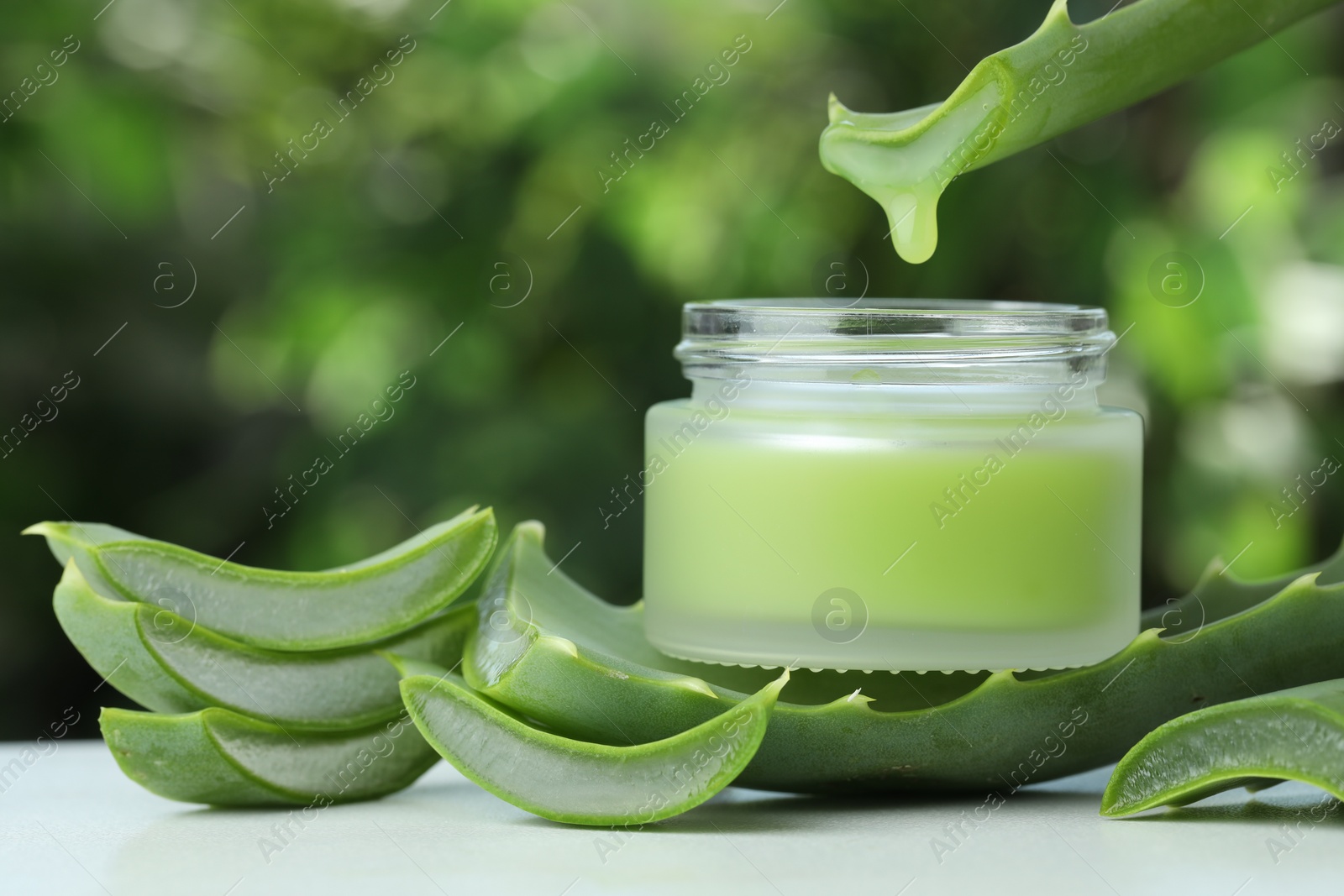 Photo of Aloe vera juice dripping from leaf into jar with cream on white table against green blurred background, closeup