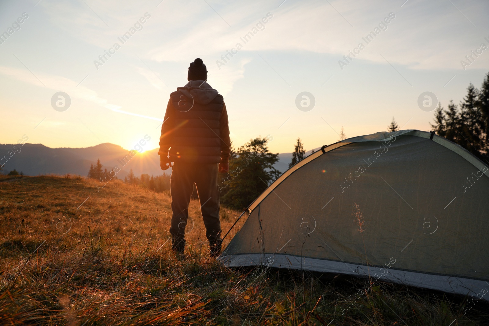 Photo of Man near camping tent in mountains at sunset, back view