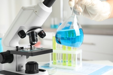 Medical assistant holding glassware with liquid near modern microscope in laboratory, closeup