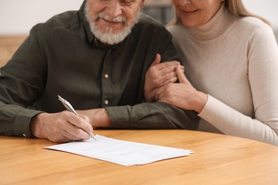 Photo of Senior couple signing Last Will and Testament at wooden table indoors, closeup