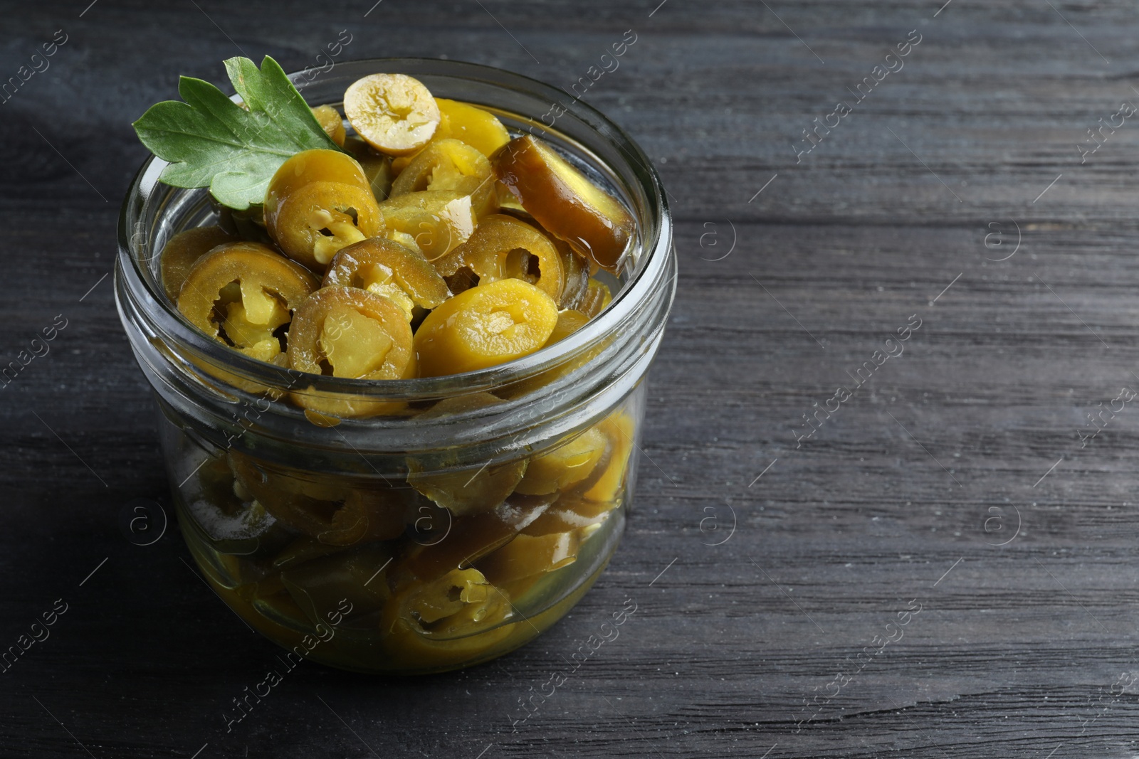 Photo of Glass jar with slices of pickled green jalapeno peppers on black wooden table, space for text