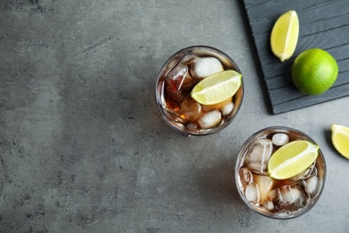 Photo of Glasses of cocktail with cola, ice and cut lime on grey background, flat lay. Space for text