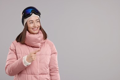 Winter sports. Happy woman with snowboard goggles pointing at something on grey background, space for text