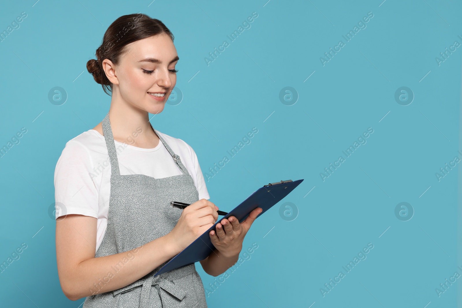 Photo of Beautiful young woman in clean apron with clipboard on light blue background. Space for text