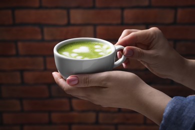 Photo of Woman with cup of delicious matcha latte against brick wall, closeup
