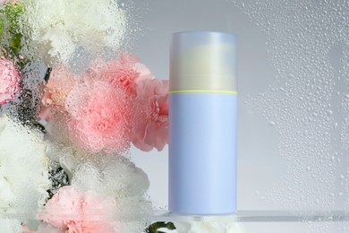 Photo of Bottle with moisturizing cream and beautiful flowers on light background, view through wet glass. Space for text