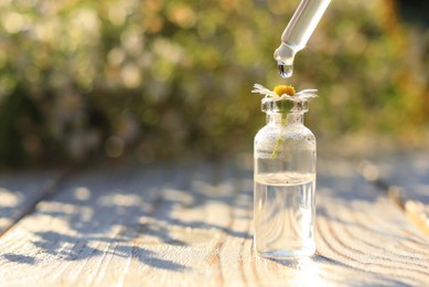 Photo of Dripping essential oil from pipette onto chamomile in bottle on white wooden table, closeup. Space for text