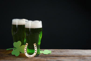 Photo of Green beer, horseshoe and clover leaves on wooden table against black background, space for text. St. Patrick's Day celebration