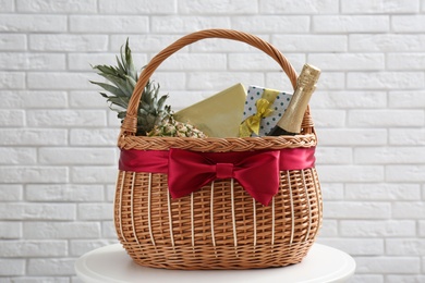 Wicker gift basket with pineapple and bottle of champagne on table near white brick wall