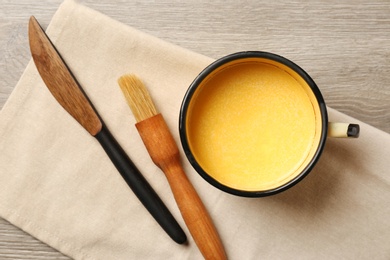 Photo of Mug of melting butter, brush and knife on wooden table, flat lay