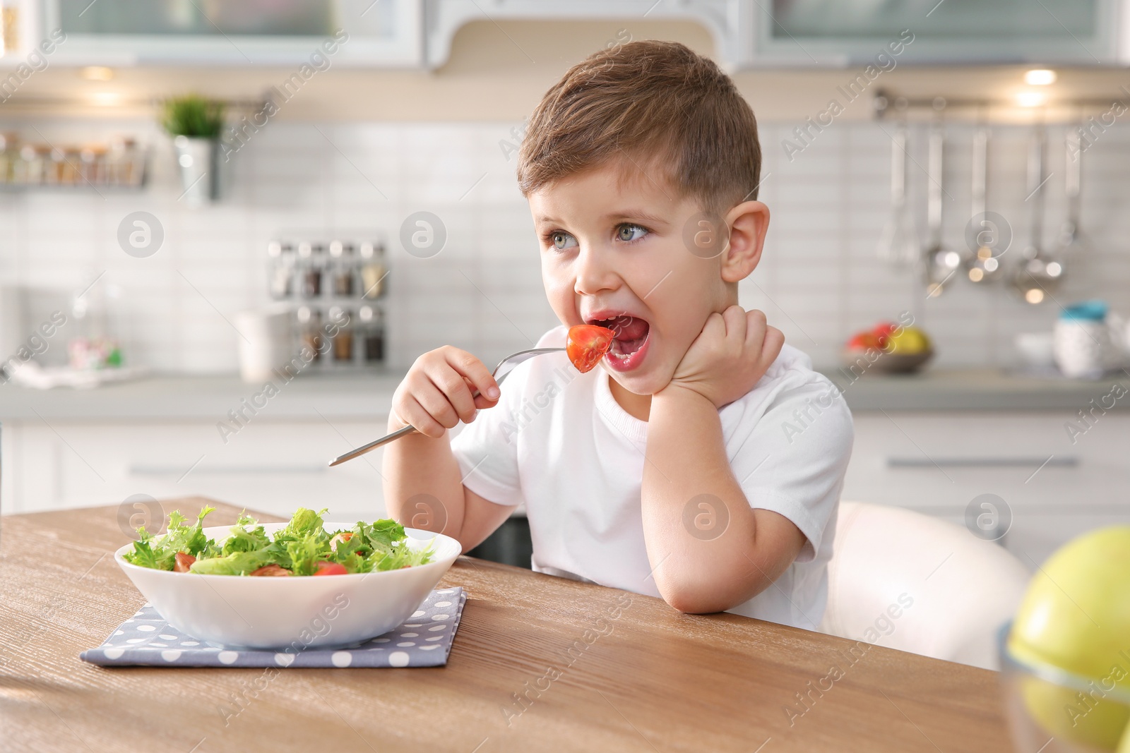 Photo of Adorable little boy eating vegetable salad at table in kitchen