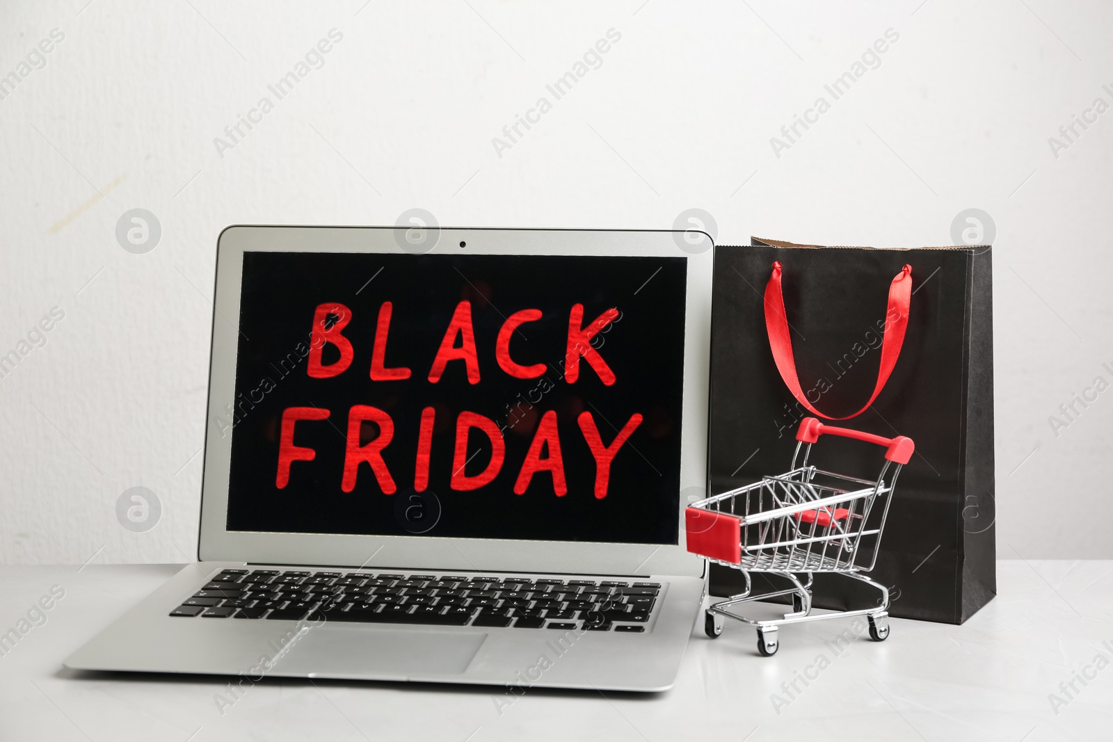 Photo of Laptop, shopping bag and small cart on grey table. Black Friday Sale online