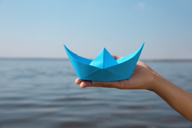 Child holding blue paper boat near river, closeup