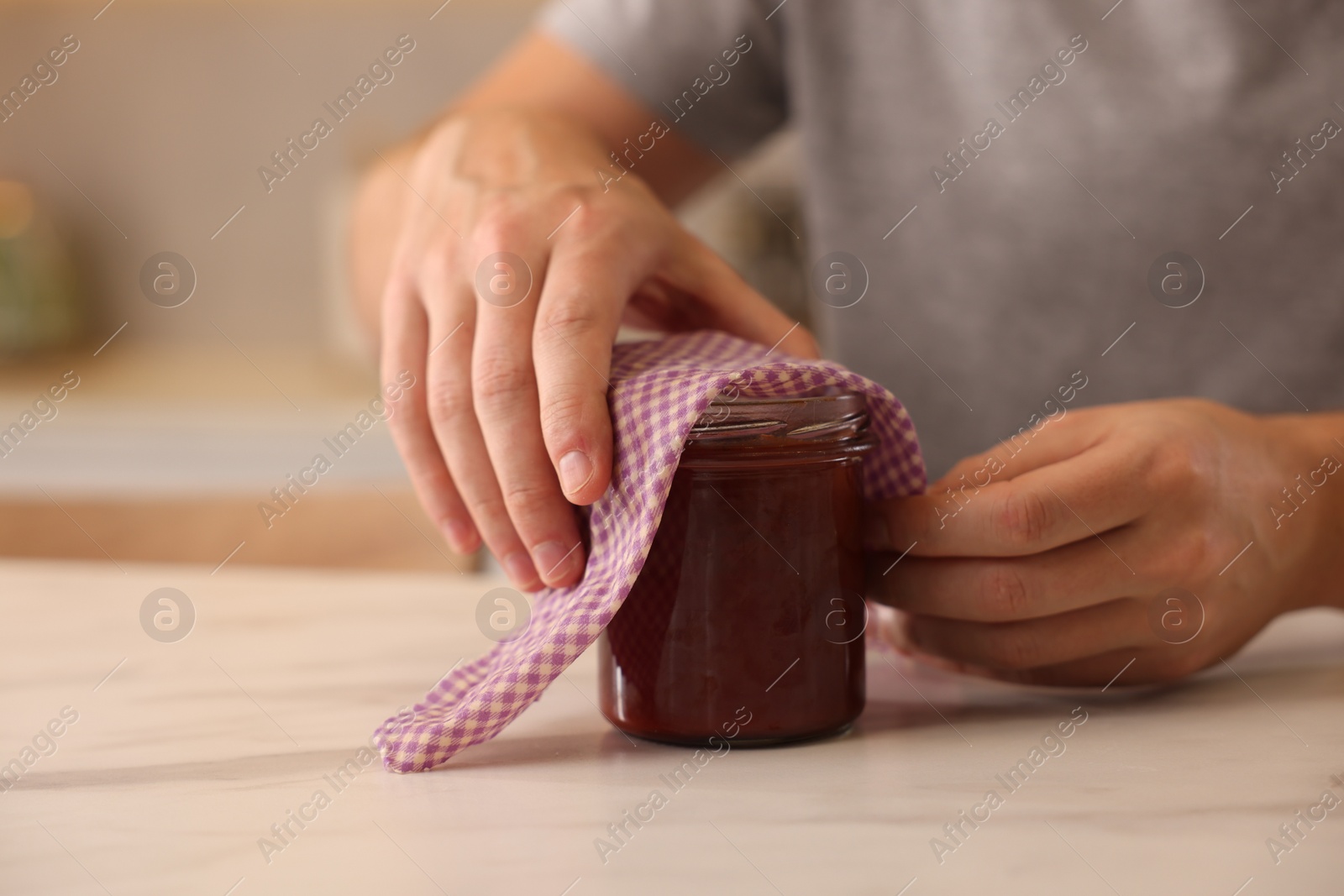 Photo of Man packing jar of jam into beeswax food wrap at light marble table indoors, closeup