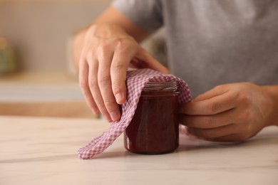 Man packing jar of jam into beeswax food wrap at light marble table indoors, closeup