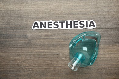 Photo of Word Anesthesia and face mask on wooden table, flat lay