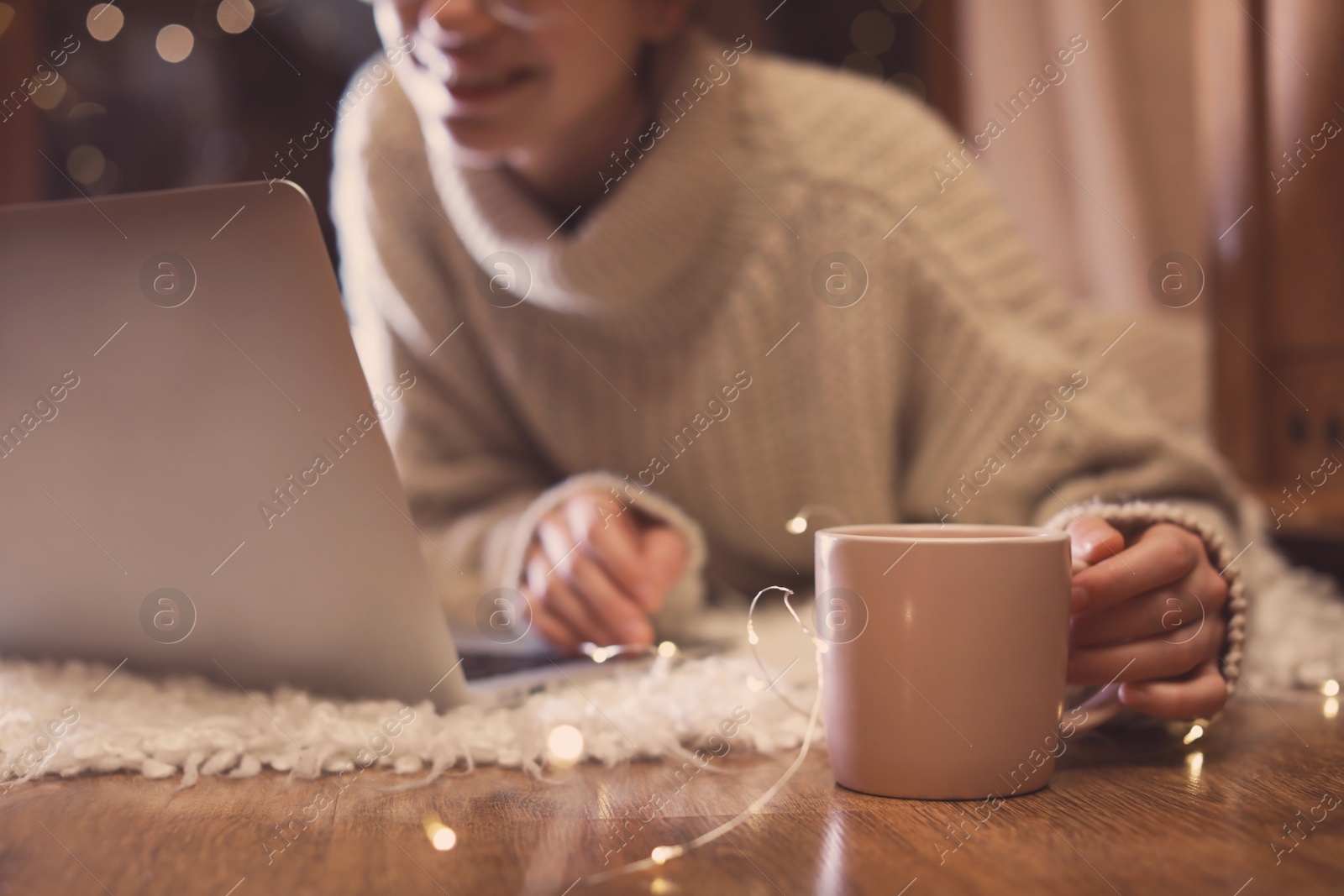 Photo of Woman with cup of hot beverage using laptop at home in winter evening, closeup