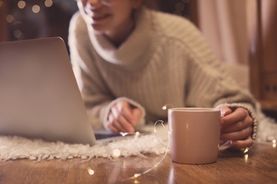 Photo of Woman with cup of hot beverage using laptop at home in winter evening, closeup