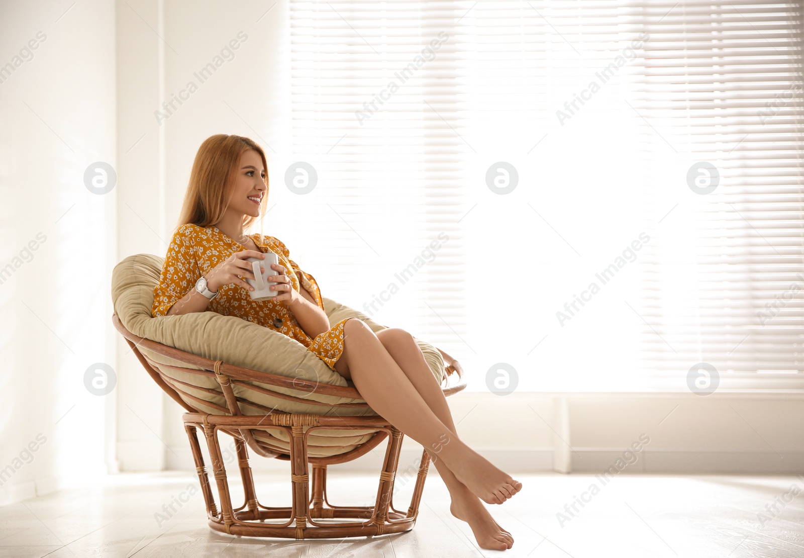 Photo of Young woman with cup of drink relaxing in papasan chair near window at home. Space for text