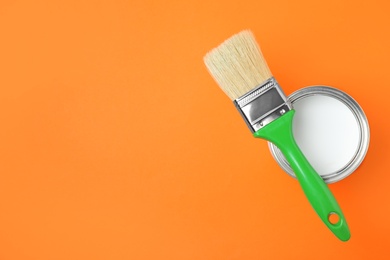 Open can with white paint and brush on orange background, top view. Space for text