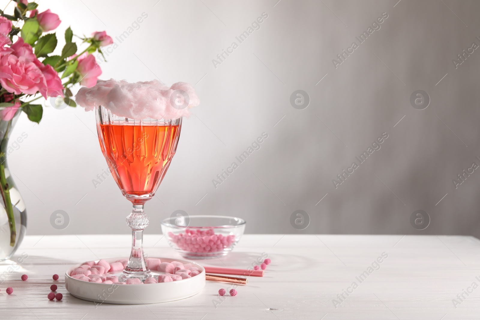 Photo of Cotton candy cocktail in glass, marshmallows, vase with pink roses and straws on white wooden table against gray background, space for text