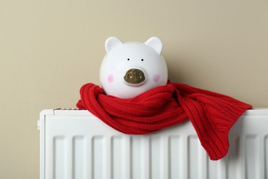 Photo of Piggy bank wrapped in scarf on heating radiator against beige background