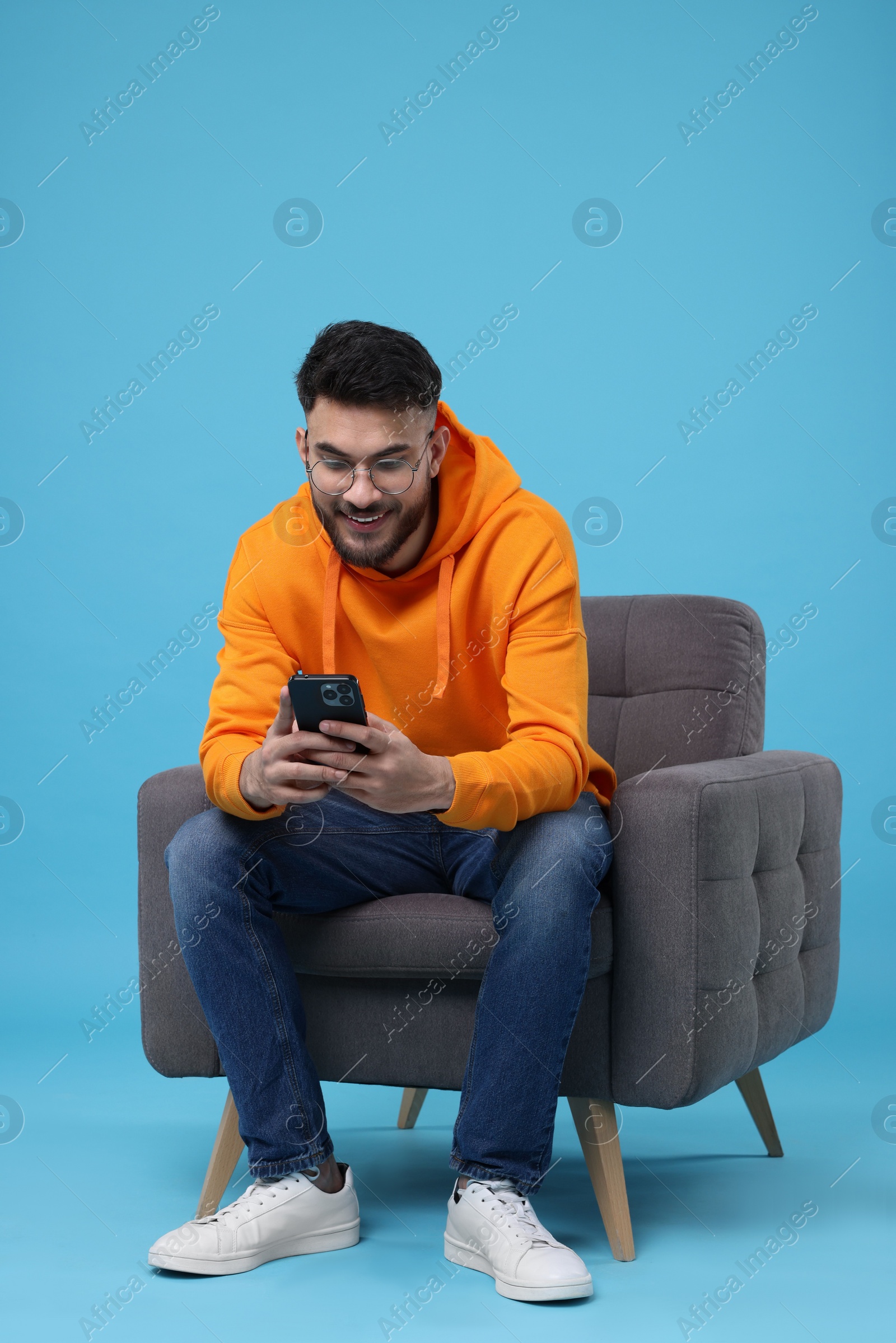 Photo of Happy young man using smartphone on armchair against light blue background