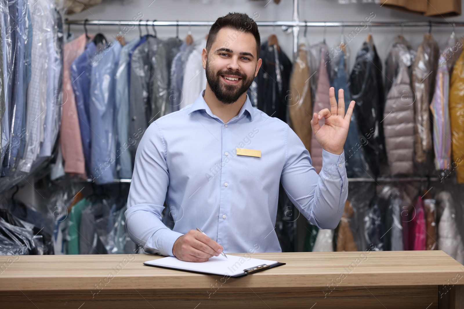 Photo of Dry-cleaning service. Happy worker showing ok gesture at counter indoors