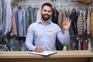 Photo of Dry-cleaning service. Happy worker showing ok gesture at counter indoors