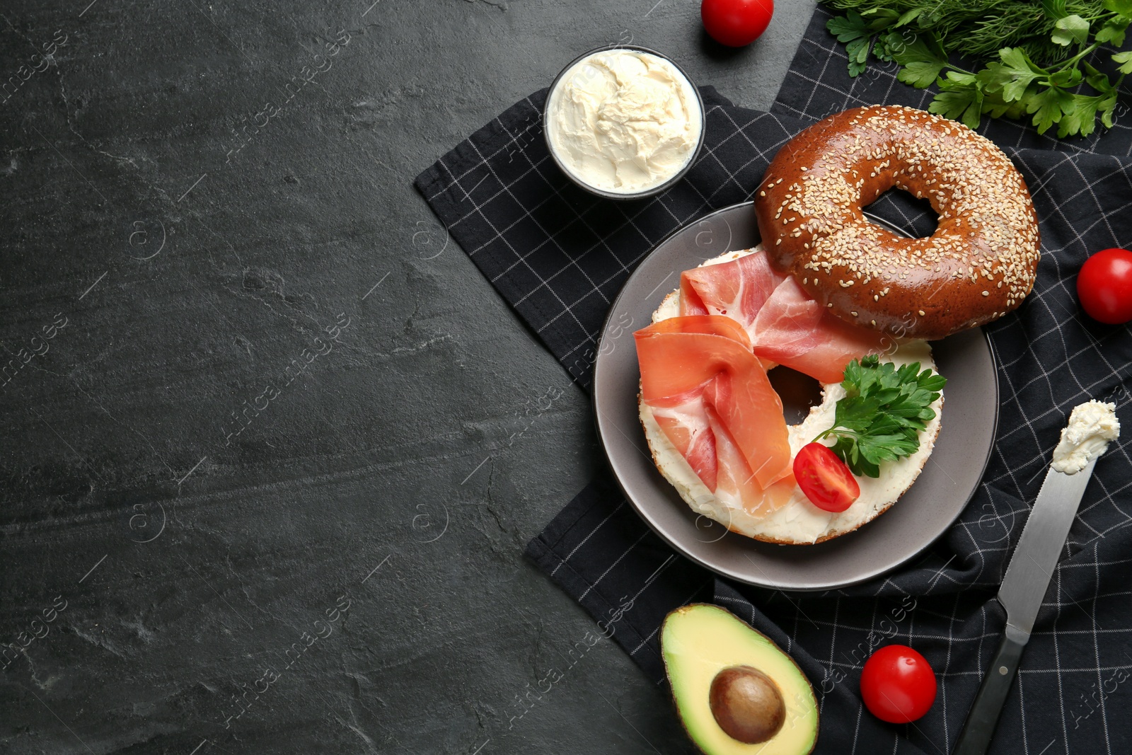Photo of Delicious bagel with cream cheese, jamon, tomato and parsley on black table, flat lay. Space for text