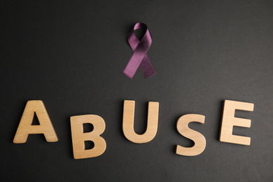 Purple ribbon and word ABUSE on black background, flat lay. Domestic violence awareness