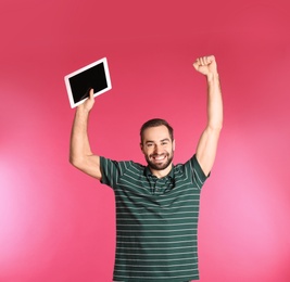 Emotional young man with tablet celebrating victory on color background