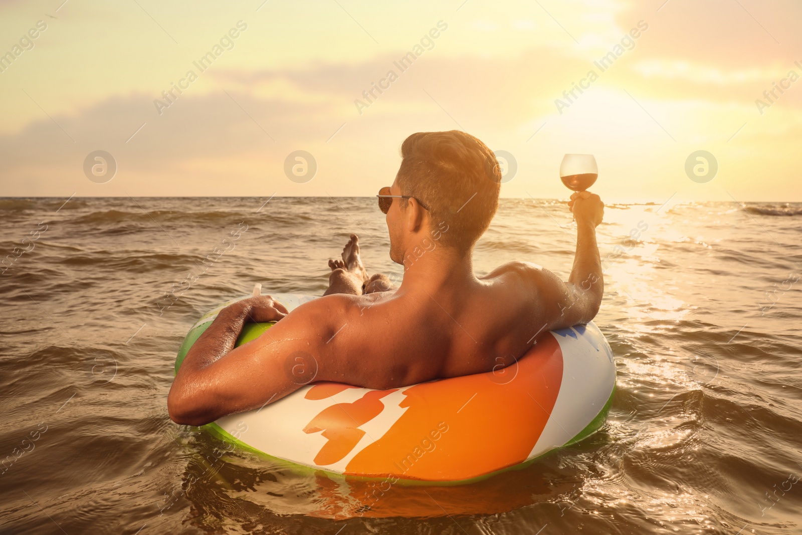 Photo of Man with glass of wine and inflatable ring resting in sea, back view