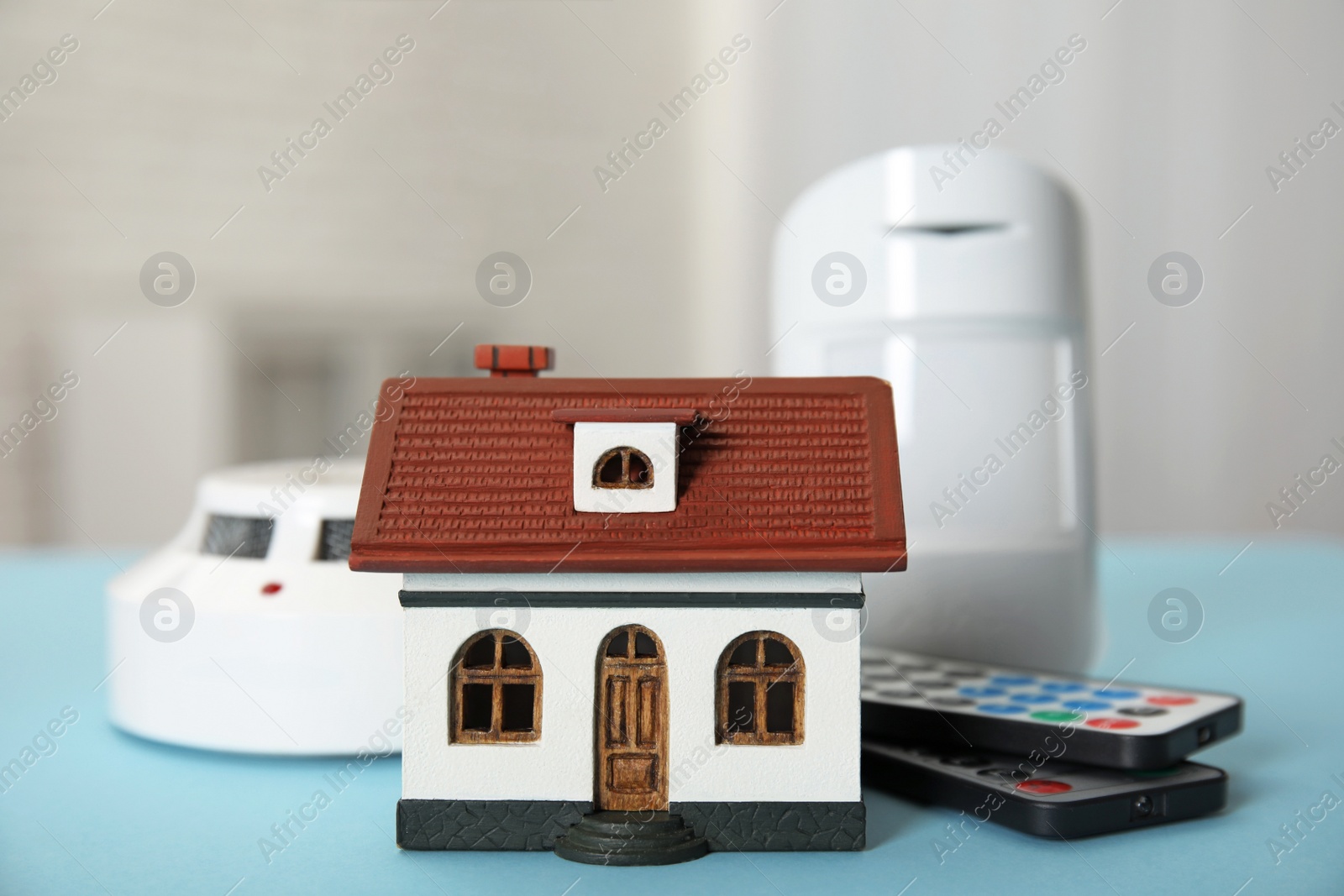 Photo of House model, remote controls, smoke and movement detectors on light blue table in room. Home security system