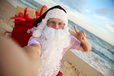 Photo of Santa Claus with bag of presents taking selfie on beach. Christmas vacation